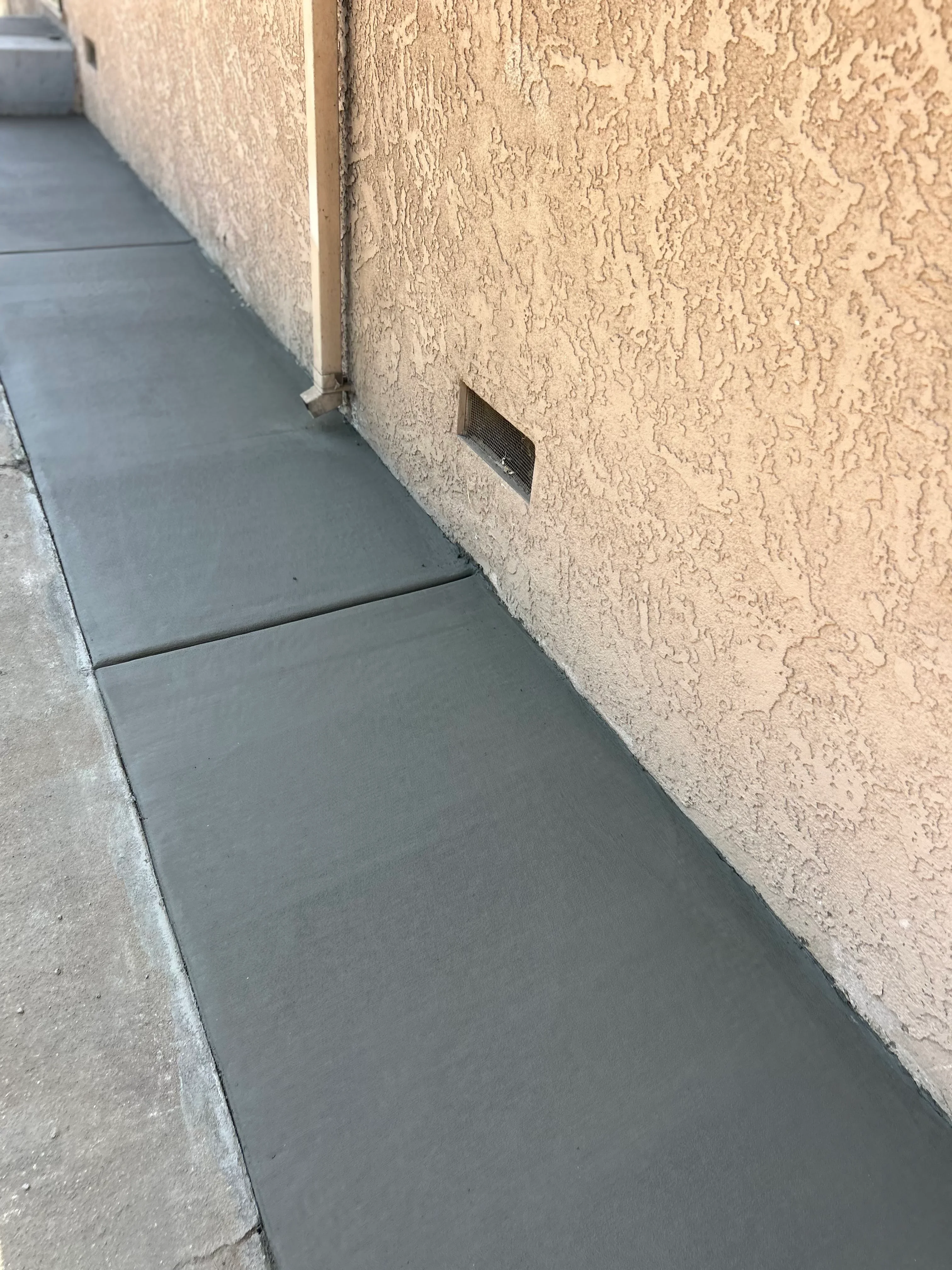 Newly installed concrete walkway with textured stucco wall and integrated vent opening